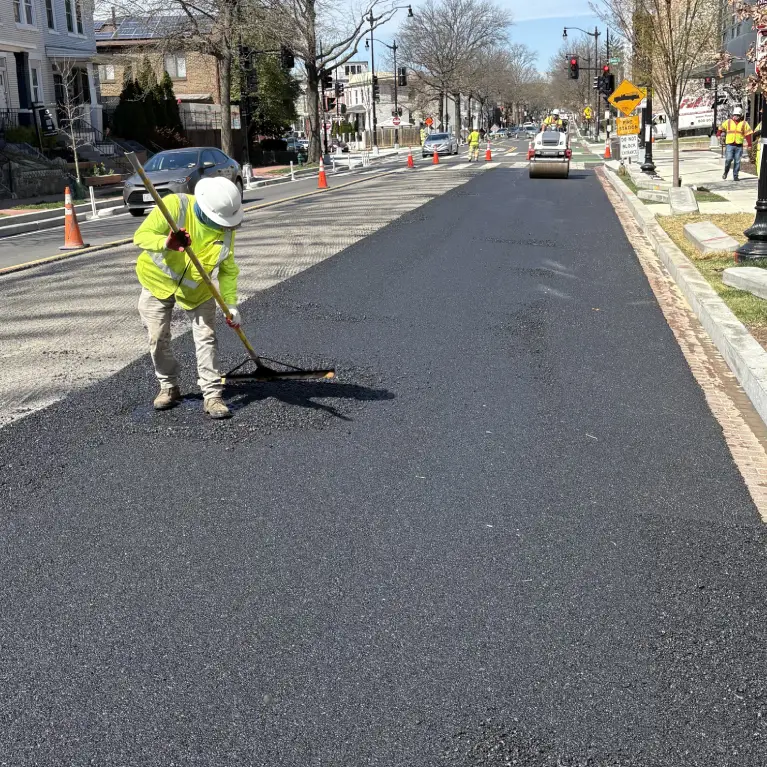A construction worker in a bright yellow safety vest and white hard hat uses a rake to spread fresh black asphalt on a city street being repaved. In the background, a steamroller and other crew members are visible.