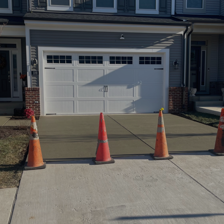 A construction worker in a bright yellow safety vest and white hard hat uses a rake to spread fresh black asphalt on a city street being repaved. In the background, a steamroller and other crew members are visible.